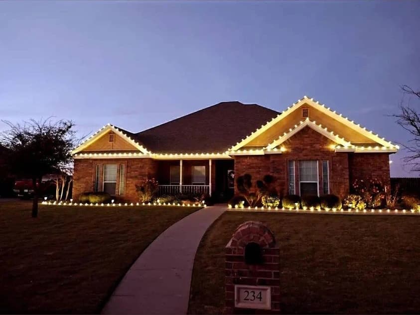 Warm-white Christmas lights outline a PLI client’s brick home and landscape borders at dusk